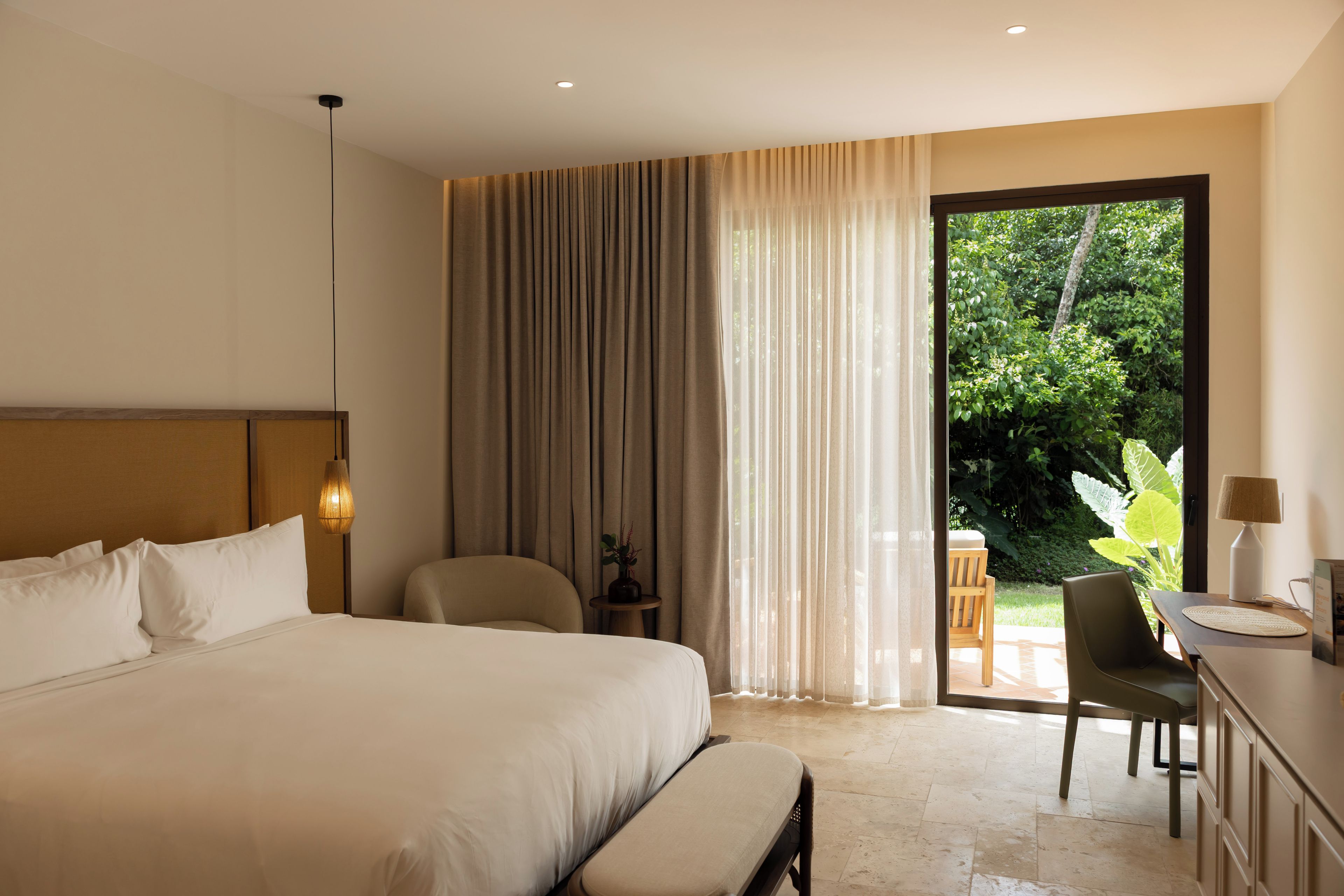 A hotel room with a large bed, a desk, a chair, a small table, and a view of greenery outside.