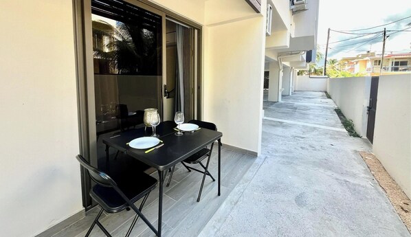 A narrow outdoor corridor with a table set for two, a chair, and a glass of water. - APT Le Sunset – Flic en Flac Air conditioning • Wi-Fi • 2 minutes from the beach (Flic en Flac)