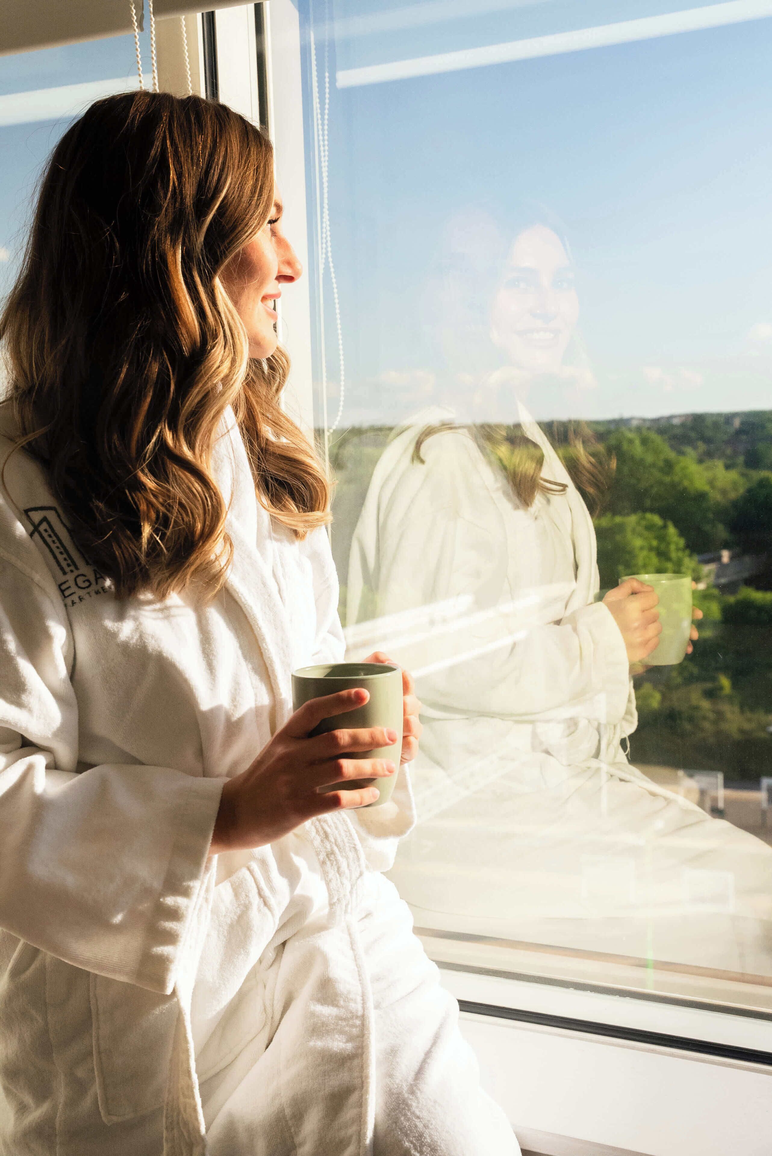 A person in a white robe sitting by a window, holding a mug.
