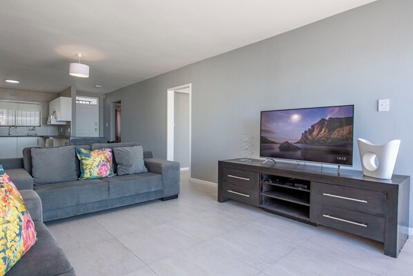 Una sala moderna con un sofá gris, un mueble de entretenimiento oscuro y un televisor de pantalla plana que muestra un paisaje de montaña.