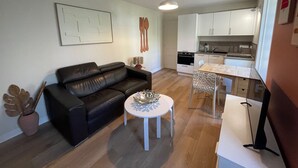 Un espace de vie compact comprenant un canapé en cuir noir, une petite table ronde, une table à manger avec des chaises, un coin cuisine et un ventilateur de plafond.