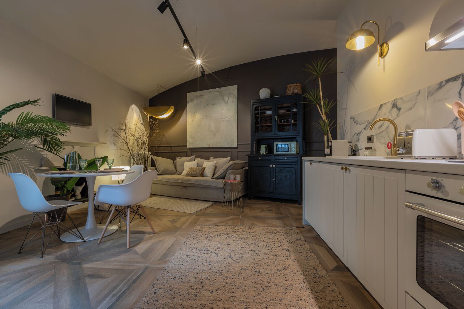 Et moderne køkken med hvide skabe, indbygget ovn og mikroovn. Der er et spiseområde med et hvidt bord og stole, samt en sofa i baggrunden.