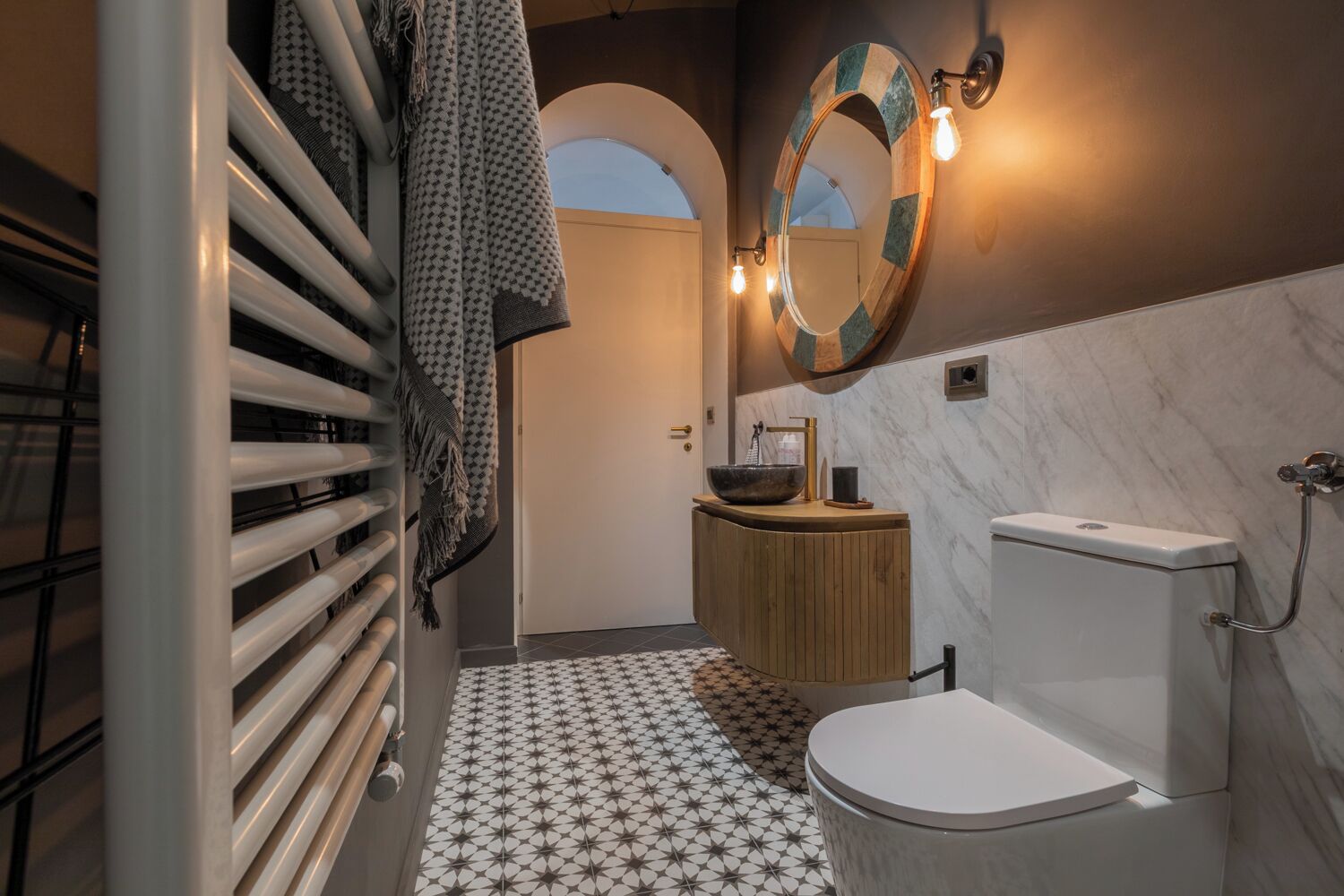 Un baño moderno con inodoro blanco, tocador de madera, espejo grande y suelo con diseño.
