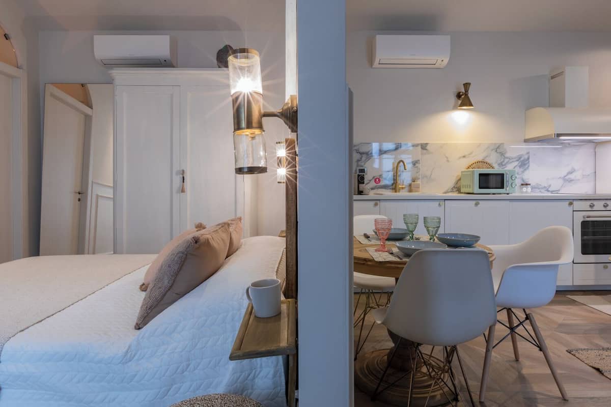 A bedroom with a bed, bedside table, and a nightstand. There is a window with curtains, a door, and an air conditioning unit on the wall.