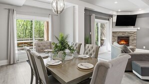 A dining area with a wooden table, upholstered chairs, a hanging light fixture, and a stone fireplace.