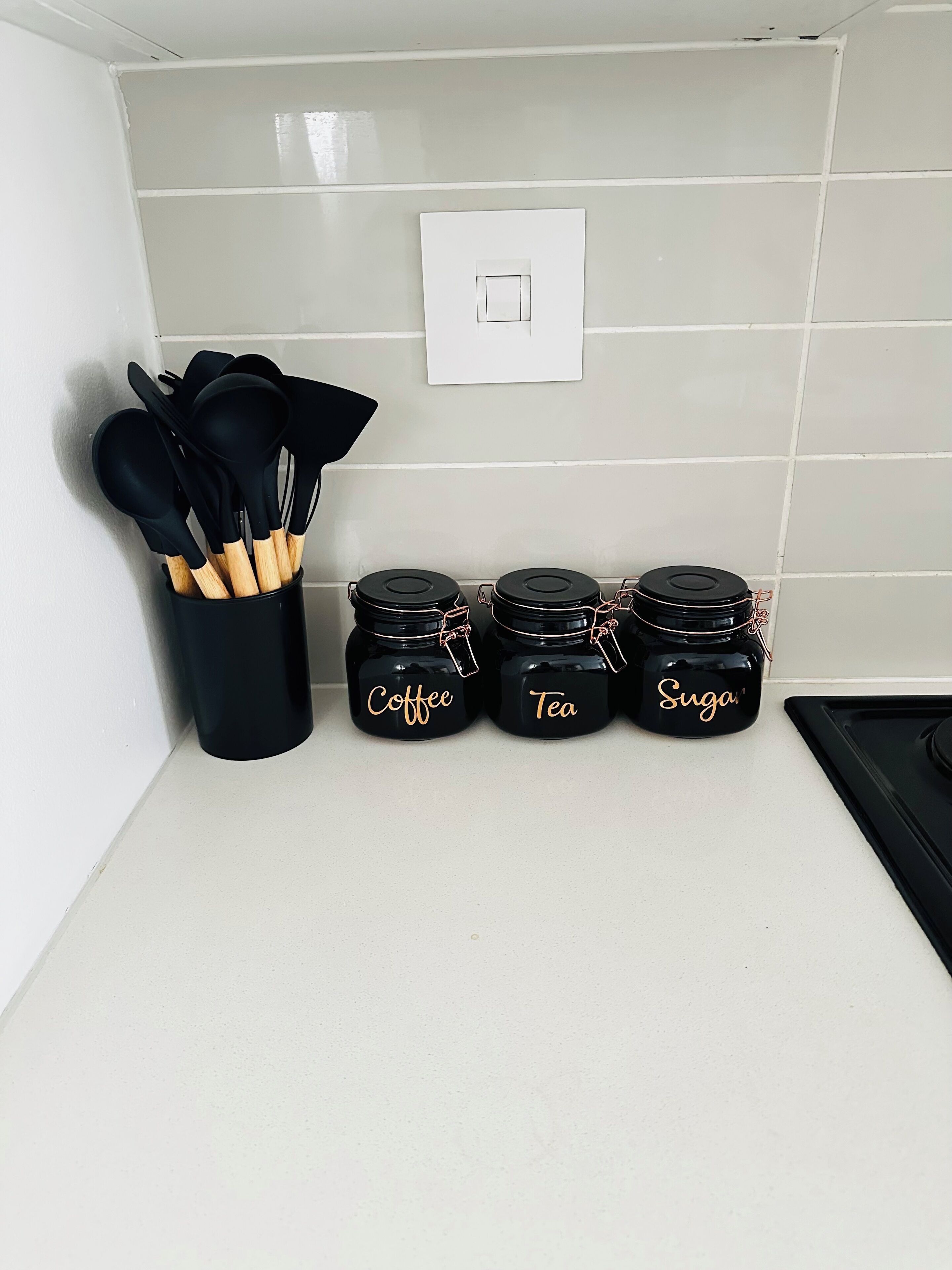Een aanrecht met een zwarte houder voor keukengerei, drie zwarte potten met de aanduiding 'Koffie', 'Thee' en 'Suiker', en een witte lichtschakelaar aan de muur.