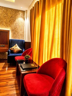 A room with a wooden floor, a blue chair with a patterned pillow, a red chair, and a table with a laptop and a plant. - 7 Hills Resort (Gwalior)