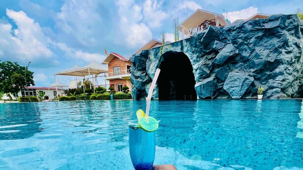 Una mano sosteniendo un vaso con pajita y una rodaja de lima, sobre una piscina azul cristalina con un fondo rocoso y una estructura similar a una cueva.