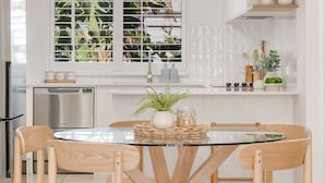 A modern kitchen with a round glass table, wooden chairs, a dishwasher, and a window with a view of greenery. - Tugun Bliss - Absolute Beachfront in Tugun - Min. 3 Night Stays - Linen Provided (Tugun)