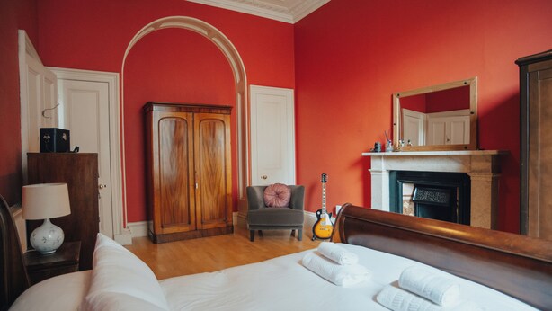 A hotel room with a large bed, a wooden wardrobe, a grey armchair, a guitar, and a fireplace. - Traditional Central 2-Bed Apartment (Edinburgh)