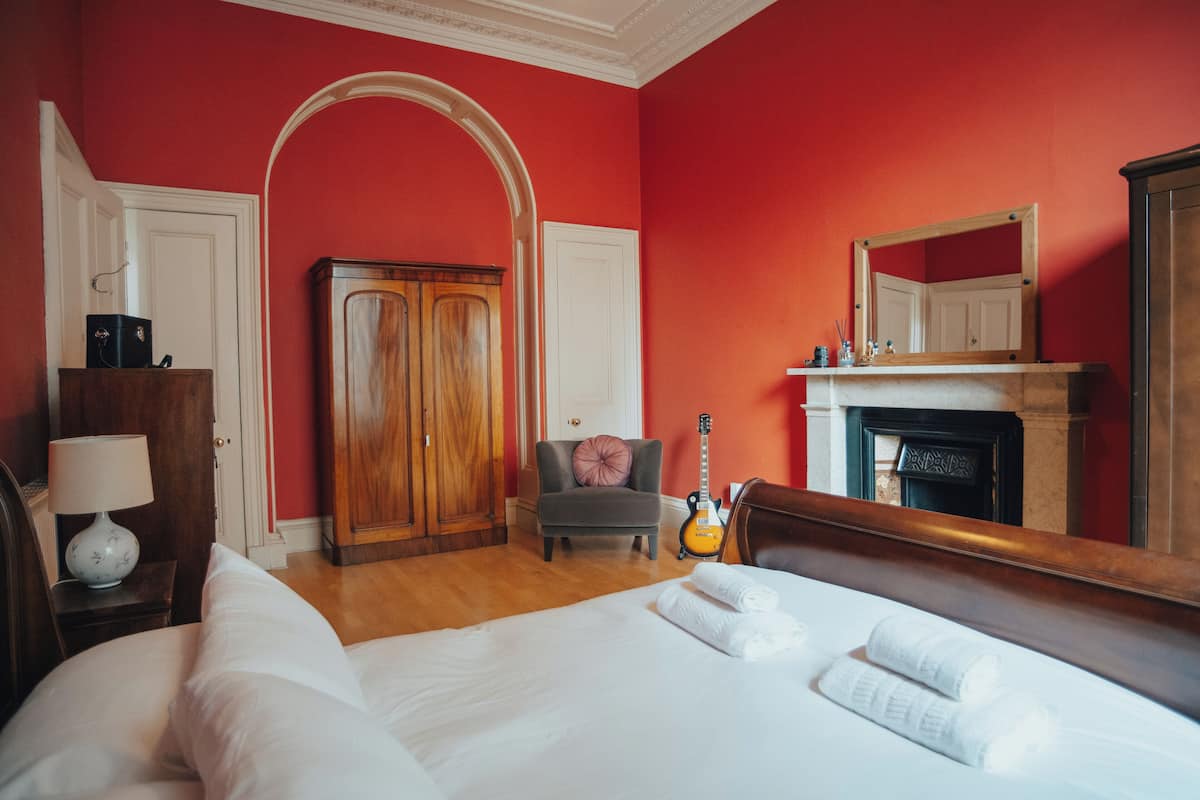 Ein Hotelzimmer mit einem großen Bett, einem Holzschrank, einem grauen Sessel, einer Gitarre und einem Kamin.