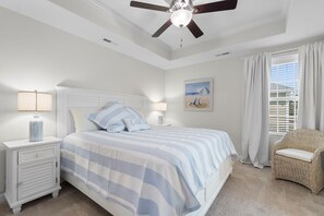 Un dormitorio con cama, mesitas de noche, ventilador de techo, una silla de mimbre y una ventana con cortinas.