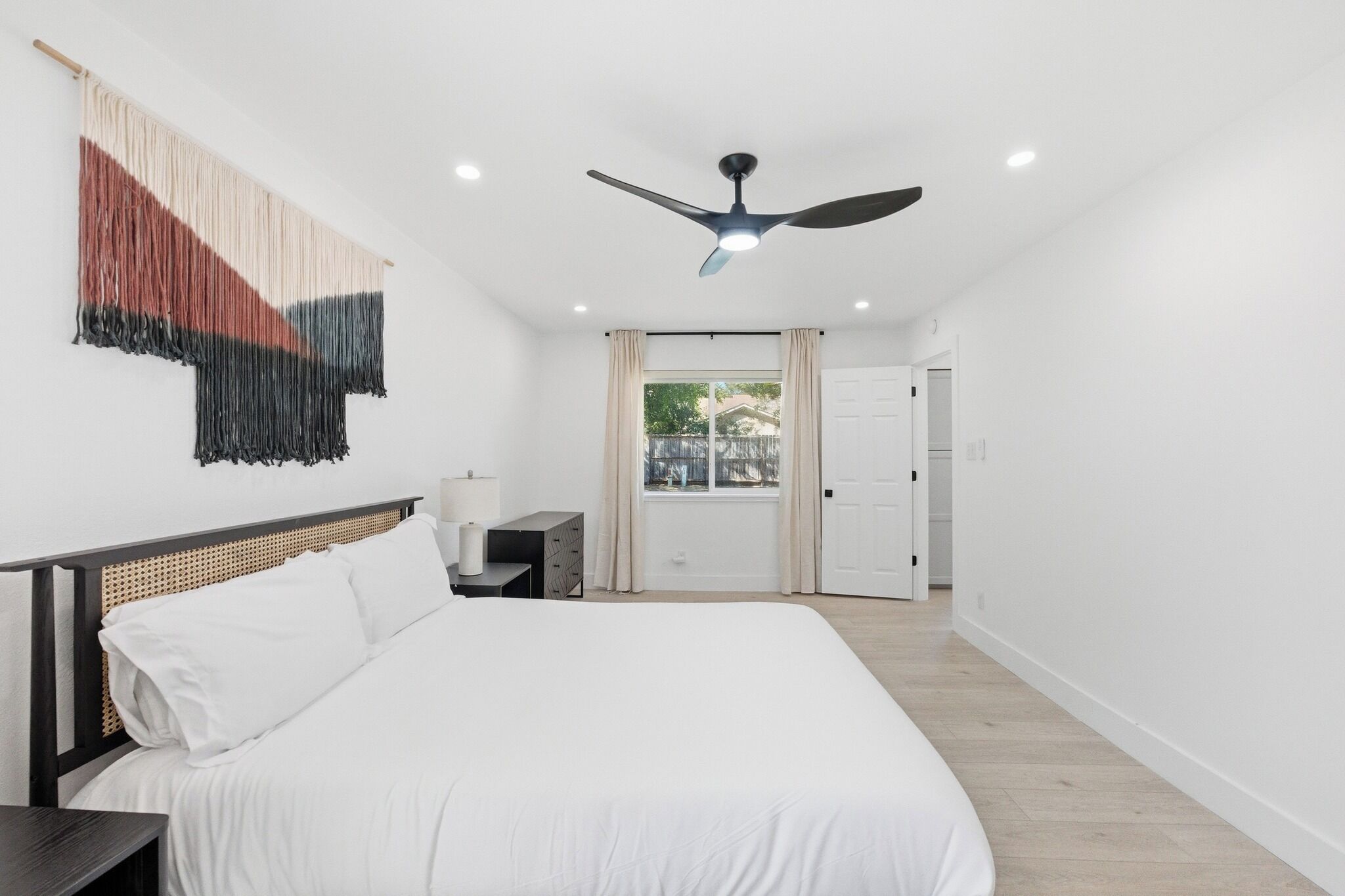 A modern bedroom with a large bed, a ceiling fan, a nightstand, and a window with curtains.
