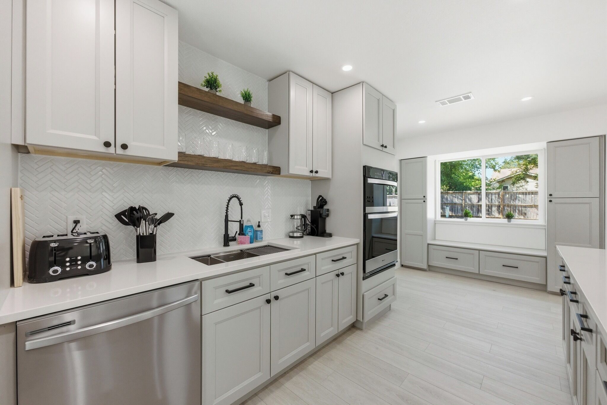 A modern kitchen with white cabinetry, a stainless steel dishwasher, a built-in oven, and a window providing a view of the outdoors.