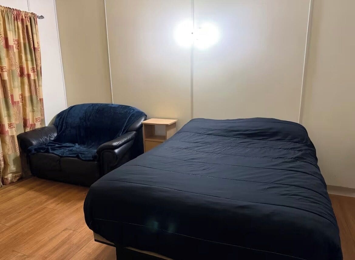 A bedroom with a bed, a chair, a bedside table, and a wall-mounted light.