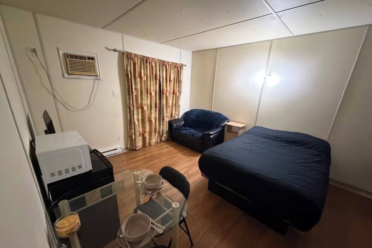 A compact hotel room with a bed, a desk with a chair, a small table with a chair, a curtain, and a wall-mounted air conditioner.