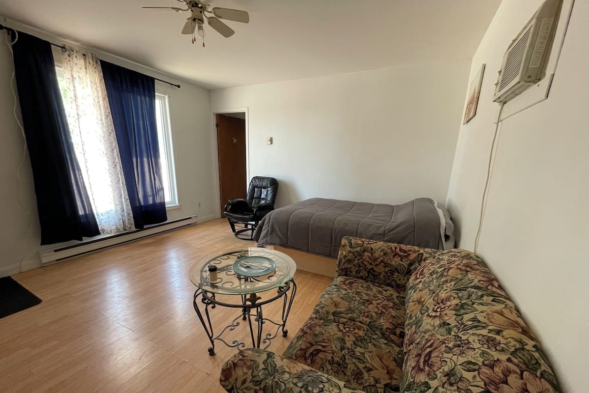 A room with a floral sofa, a glass coffee table, a leather chair, a bed, and a ceiling fan.