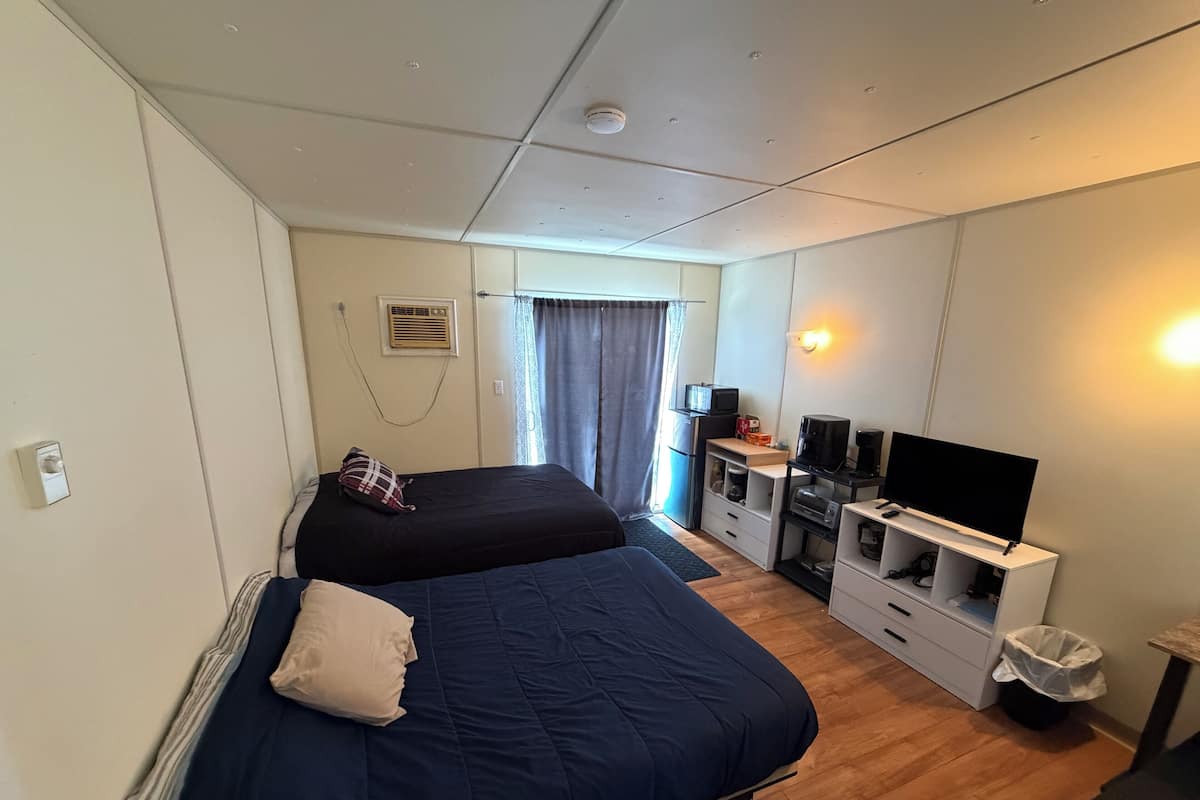 A compact hotel room with two beds, a desk with a computer, a TV, and a small kitchenette.