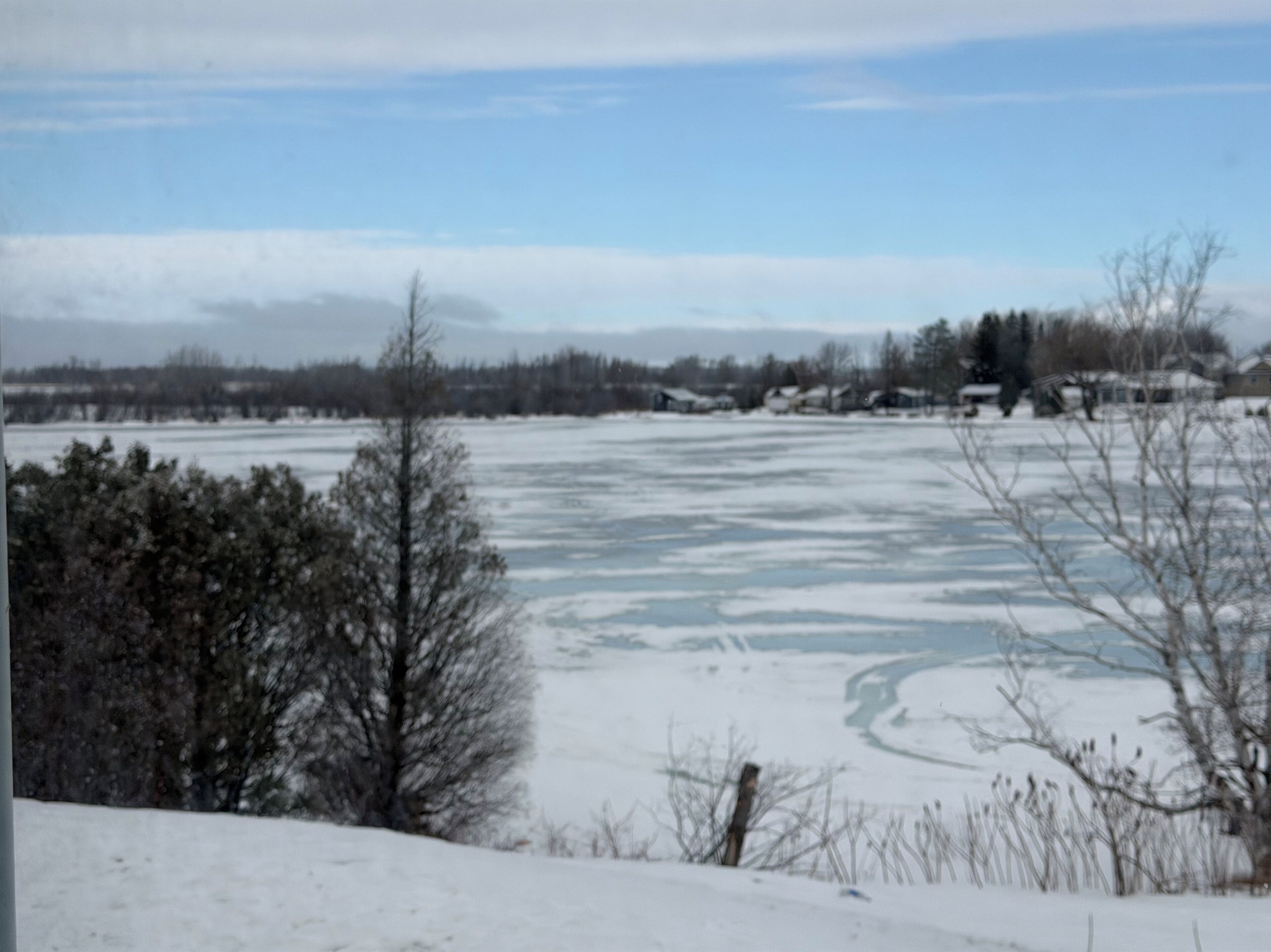Paesaggio innevato con un lago gelato, alberi e case sullo sfondo.