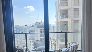 Ein Balkon mit Korbstühlen und einem Tisch, mit Blick auf eine Stadtlandschaft mit Gebäuden und Baukränen.