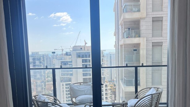 Un balcon avec des fauteuils en osier et une table, d’où l’on peut admirer un panorama urbain avec des immeubles et des grues de construction.