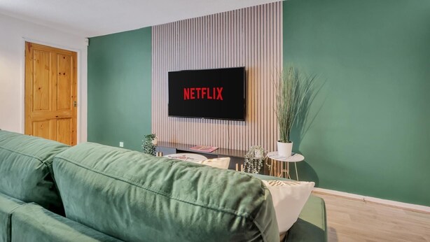 Un salón con un sofá verde, una puerta de madera, un televisor colocado en una pared a rayas y una mesita con una planta.