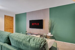 Ein Wohnzimmer mit einem grünen Sofa, einer Holztür, einem an einer gestreiften Wand montierten Fernseher und einem kleinen Tisch mit einer Pflanze.