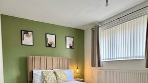 Un dormitorio con cama, paredes verdes, cuadros enmarcados, un radiador y una ventana con cortinas.