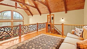 A spacious loft with a high wooden ceiling, a large window offering a view of trees, a staircase with a wooden railing, and a cozy seating area with a patterned rug.
