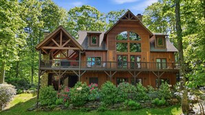 A large wooden house with a balcony, surrounded by lush greenery and a stone wall.