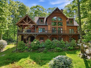 A large wooden house with a balcony, surrounded by lush greenery and a stone wall. - 5BR, 4000 sq ft Luxury Cabin w/ Covered Fireplace! (Blowing Rock)