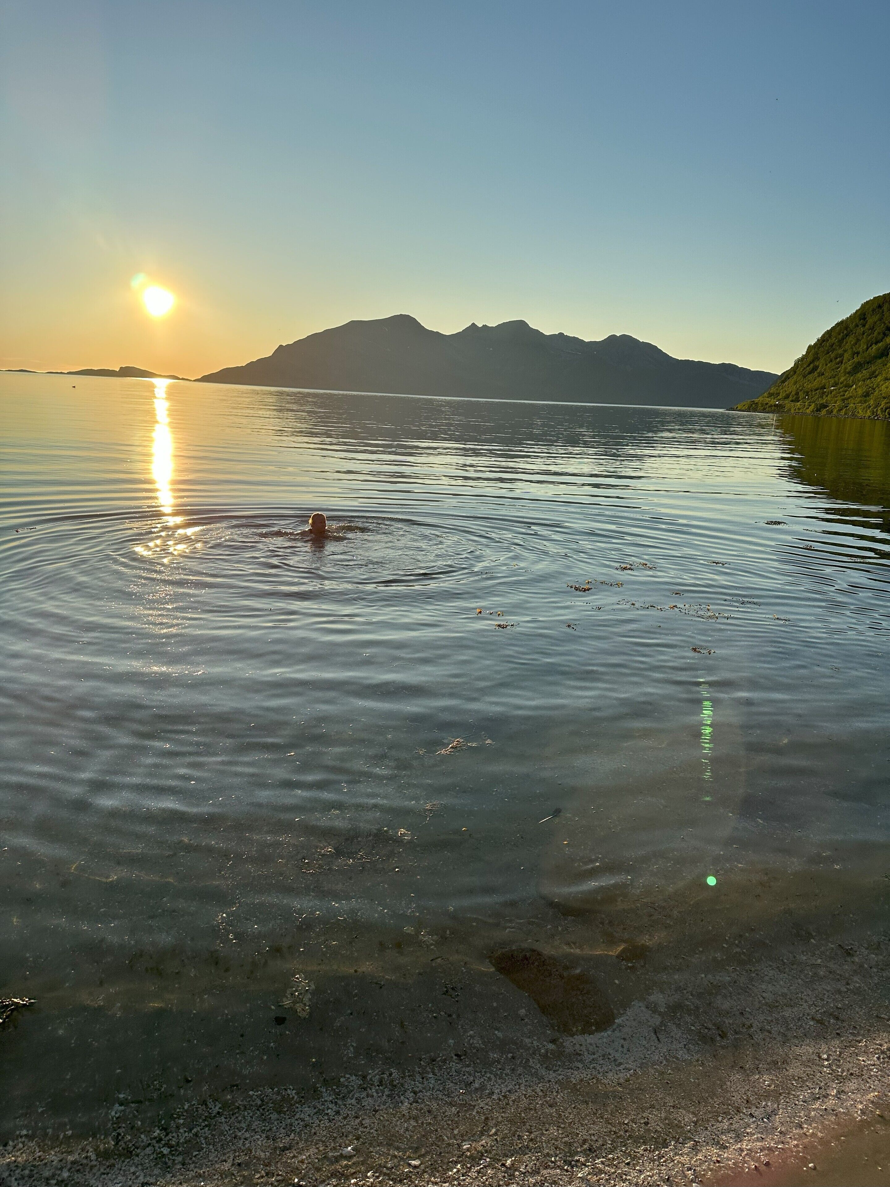 Ein ruhiger See mit einem klaren Spiegelbild der Sonne und der Berge im Hintergrund.