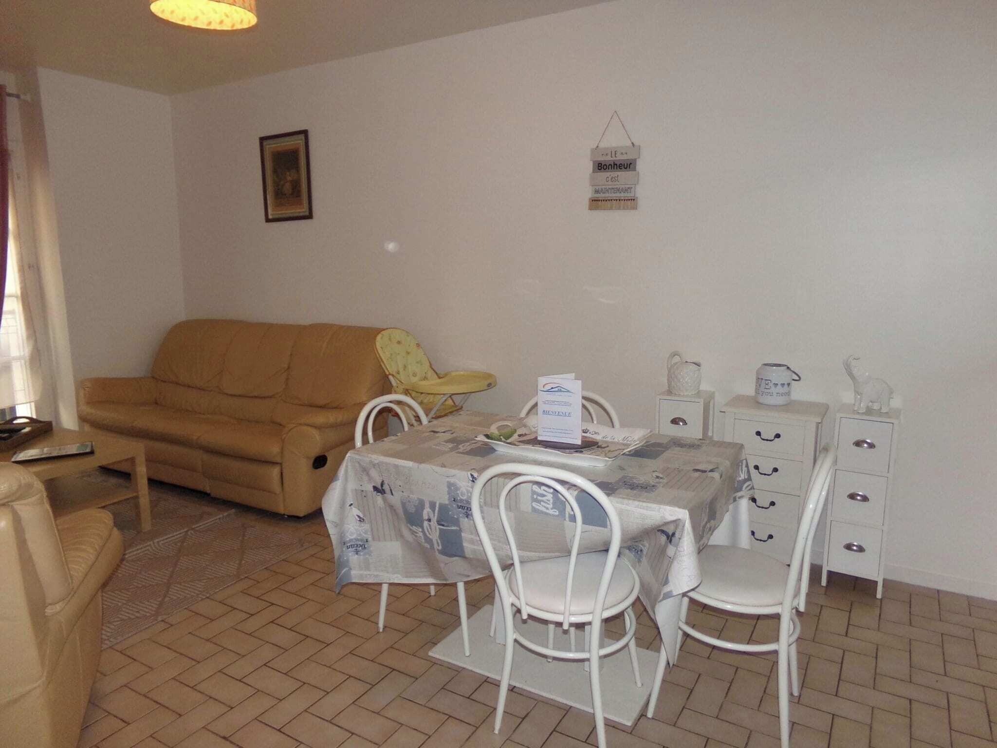 Ein Wohnzimmer mit einer Couch, einem Esstisch mit Stühlen, einem kleinen Tisch mit Wasserkocher und Tassen sowie einem begrüßungshinweis im Bilderrahmen an der Wand.