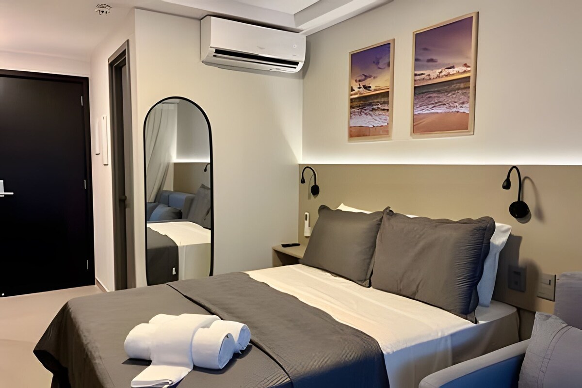 A hotel room with a bed, a bedside table, a mirror, an air conditioning unit, and two framed pictures on the wall.
