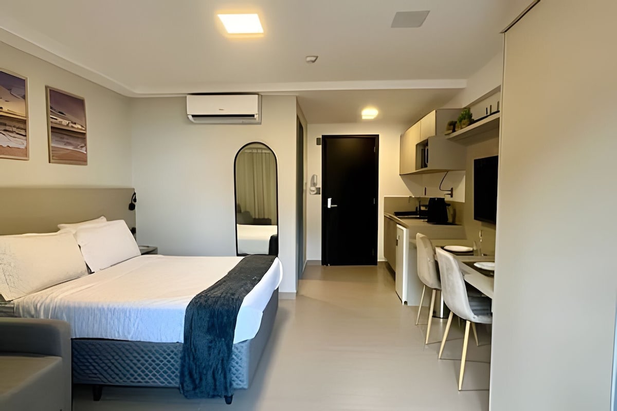 A modern hotel room with a bed, bedside table, mirror, air conditioning unit, and a kitchenette with a dining area.