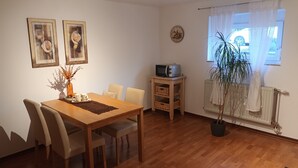 A dining area with a wooden table and chairs, a small TV on a stand, a potted plant, and a window with curtains. - Holiday Apartment Doris, 57sqm, 1 Living-/Bedroom, max 2 People (Sinsheim)