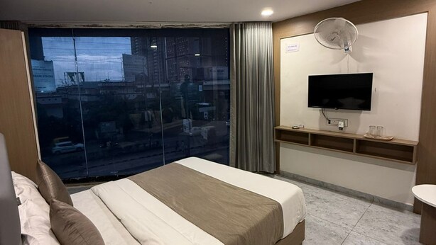 A modern hotel room with a large bed, a TV, a fan, and a view of the city through a large window. - Hotel Urban View (Ahmedabad)