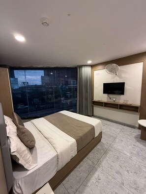 A modern hotel room with a large bed, a TV, a fan, and a view of the city through a large window. - Hotel Urban View (Ahmedabad)