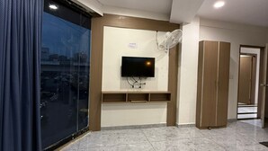 A modern living room with a flat-screen TV mounted on a wall, a ceiling fan, and a sliding glass door leading to a balcony. - Hotel Urban View (Ahmedabad)