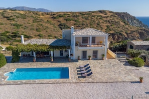 Una casa de dos pisos con techo de piedra, piscina y un patio con sillones de descanso.