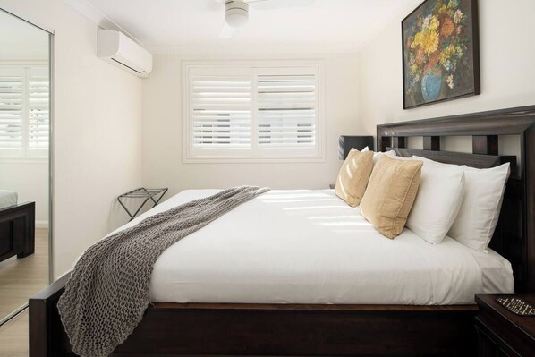 A neatly made bed with white linens and two beige pillows, a dark wooden headboard, a small bedside table, and a wall-mounted air conditioner.