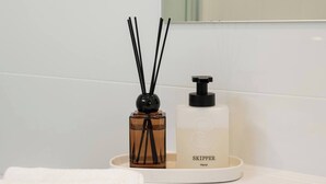Une salle de bain avec un lavabo blanc, une serviette blanche, un diffuseur d’huiles essentielles en roseau marron et un distributeur de savon liquide blanc.