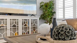 Un salón con televisor mostrando una escena de playa, un mueble blanco, una planta y una cesta decorativa con diversos objetos.