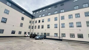 Un immeuble à plusieurs étages, doté d’une façade sobre, de nombreuses fenêtres et d’un espace de stationnement où sont garées plusieurs voitures.