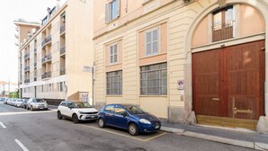 Straßenansicht eines Wohnhauses mit mehreren Balkonen, einem parkenden Auto und einer großen Holztür.