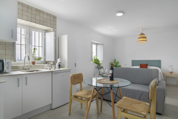 A modern kitchen with white cabinets, a dining table with chairs, a sofa, and a bed with a headboard.