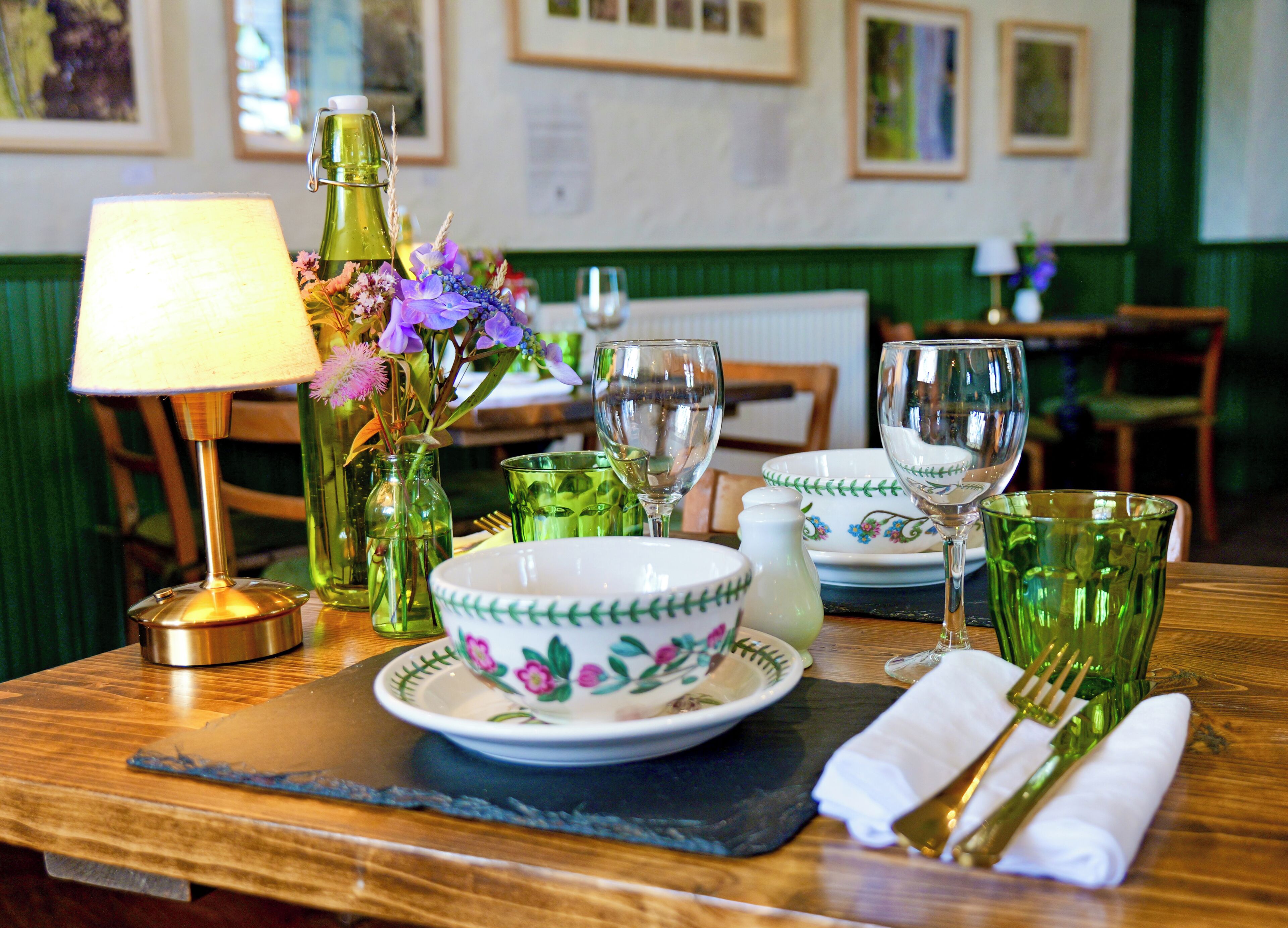 A dining table set with a bowl, plates, glasses, a lamp, and a vase with flowers.