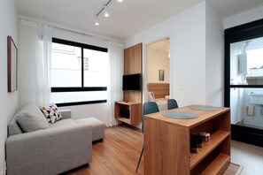 A modern hotel room with a sofa, a wooden dining table, a TV, and a bathroom visible through an open door. - Boulevard Agami (São Paulo)