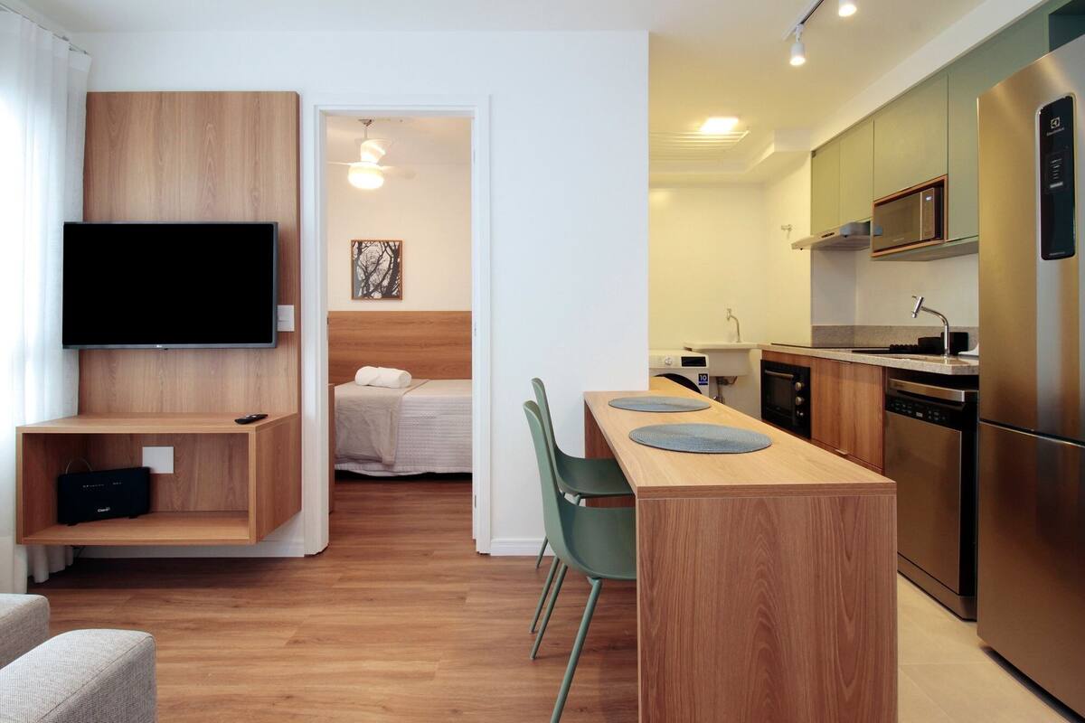 A modern, compact living space with a wooden accent wall, a flat-screen TV, a bed, a kitchenette, and a dining area.
