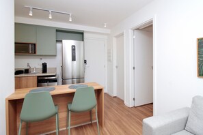 A modern kitchen with stainless steel appliances, a wooden dining table with two chairs, and a grey sofa in a bright room with white walls and wooden flooring. - Boulevard Agami (São Paulo)